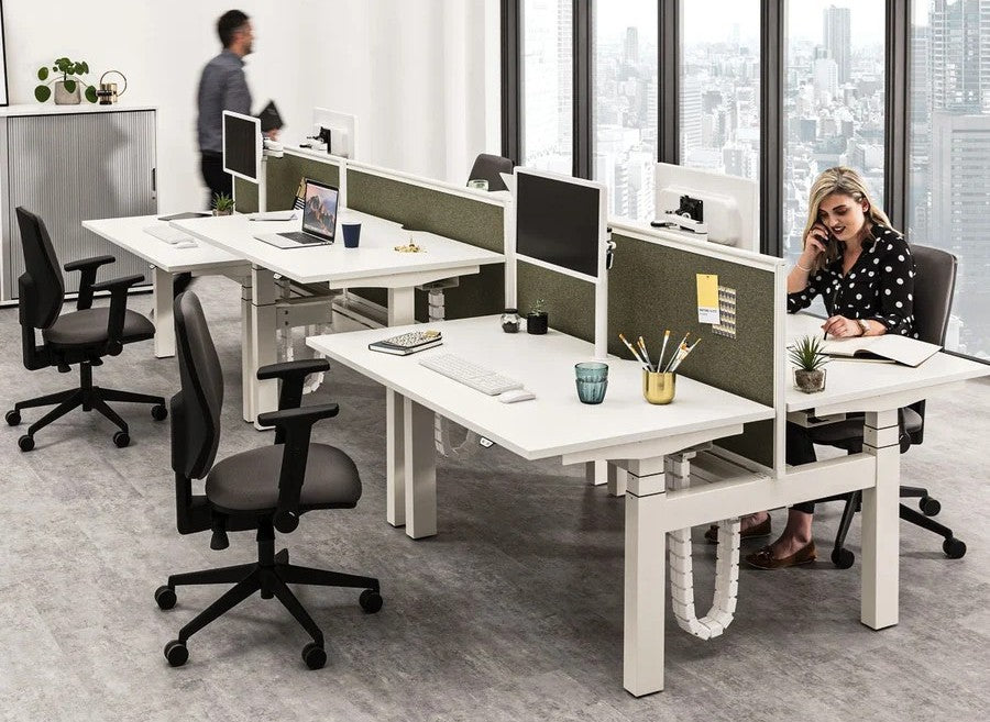 An office with two rows of modern, back-to-back height-adjustable desks with white tops and fabric divider screens, featuring a person sitting and working at one desk.