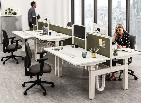 An office with two rows of modern, back-to-back height-adjustable desks with white tops and fabric divider screens, featuring a person sitting and working at one desk.